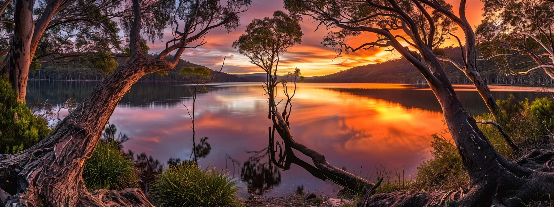a vibrant sunset casts dramatic shadows across a tranquil lake, with lush trees framing the serene water reflecting the fiery sky.